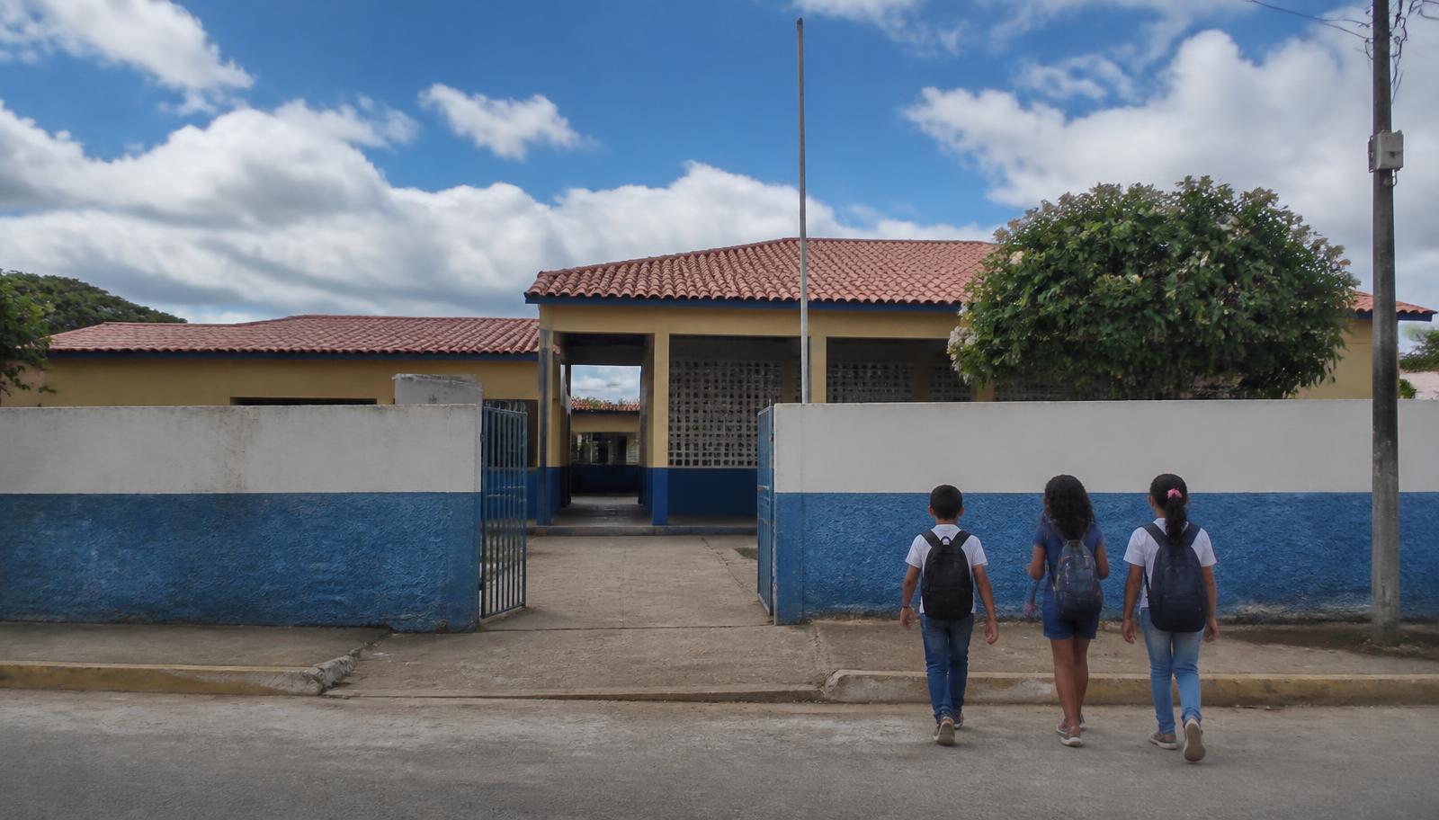 Fachada de escola municipal no interior do Nordeste com crianças chegando de mochila